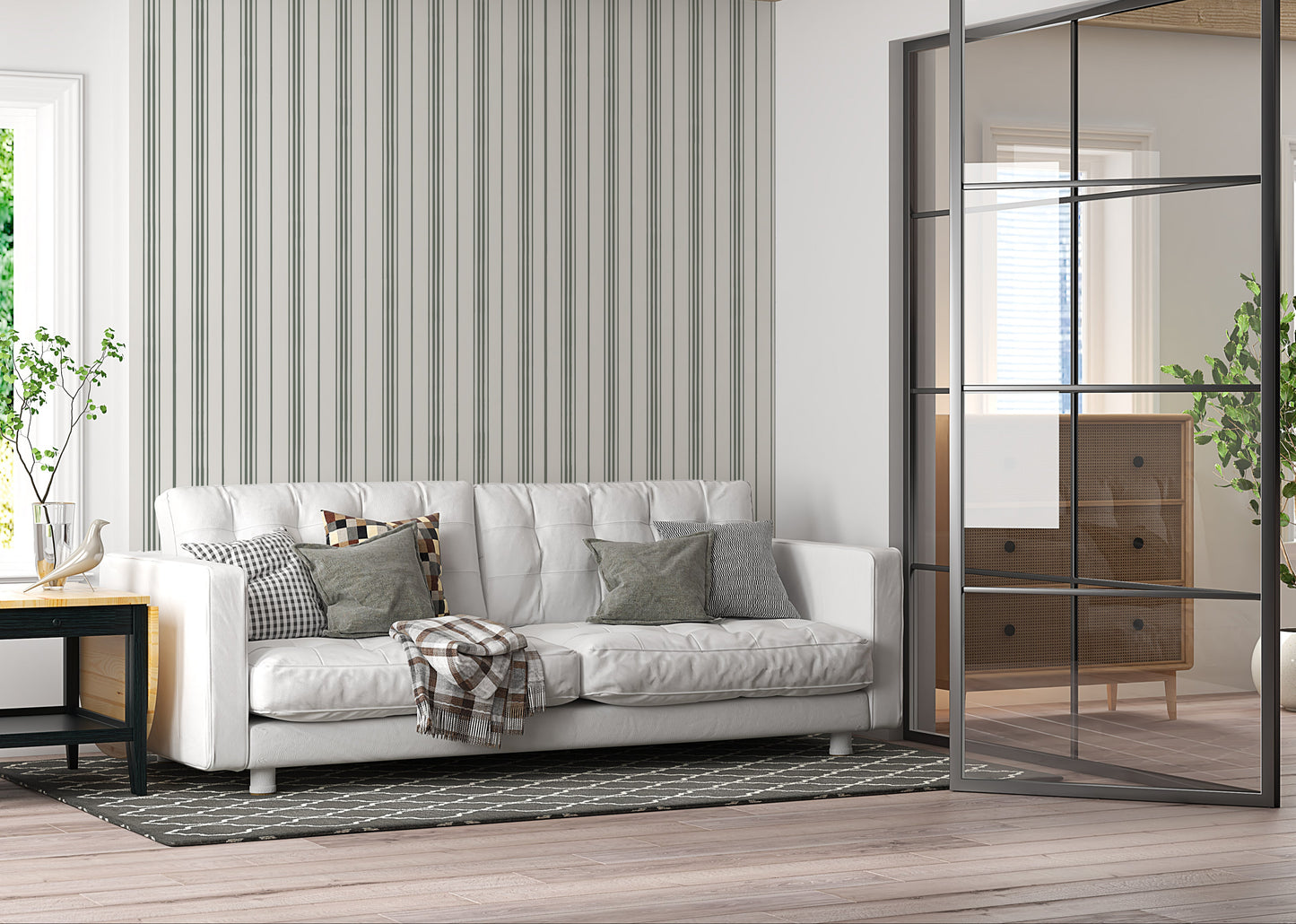 An airy, modern living room featuring a white tufted sofa against a wall with vertical grey and olive green stripes. The sofa is adorned with a mix of plaid, checkered, and solid olive-colored throw pillows, along with a draped plaid blanket. It sits on a dark grey patterned area rug over light-colored hardwood floors. To the left is a small black and light wood side table holding a vase and a bird figurine. On the right, a large, black metal-framed glass sliding partition separates the space from another 