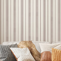 A close-up shot of a beige couch layered with an assortment of textured throw pillows, set against a backdrop of cream-colored wallpaper with vertical brown stripes. The pillows feature various designs, including a grey chunky knit, a white and grey geometric diamond pattern, a smooth brown leather accents, and a bright mustard yellow knit. A fluffy brown faux-fur blanket is also draped over the back of the sofa, creating a warm, cozy interior aesthetic.