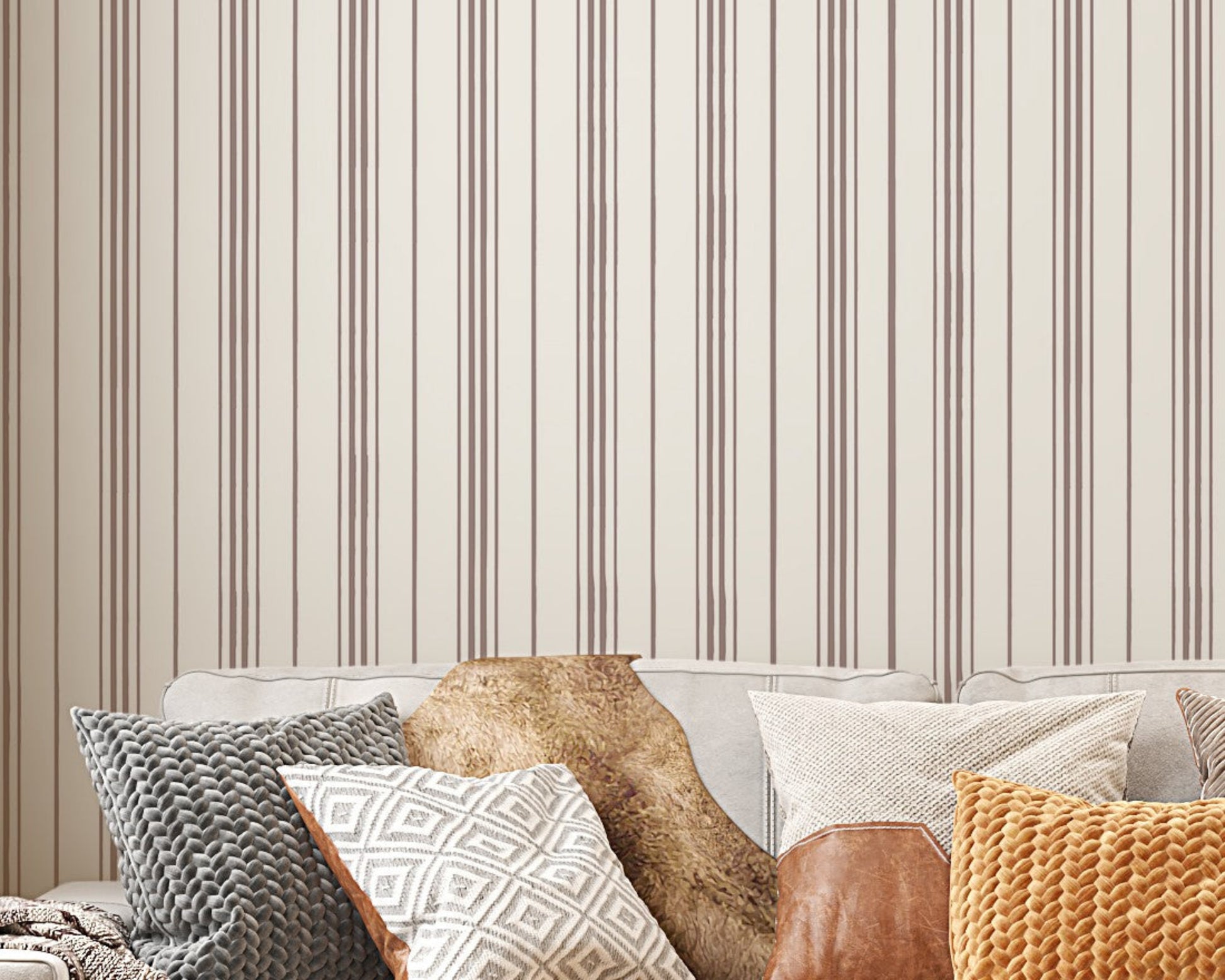 A close-up shot of a beige couch layered with an assortment of textured throw pillows, set against a backdrop of cream-colored wallpaper with vertical brown stripes. The pillows feature various designs, including a grey chunky knit, a white and grey geometric diamond pattern, a smooth brown leather accents, and a bright mustard yellow knit. A fluffy brown faux-fur blanket is also draped over the back of the sofa, creating a warm, cozy interior aesthetic.