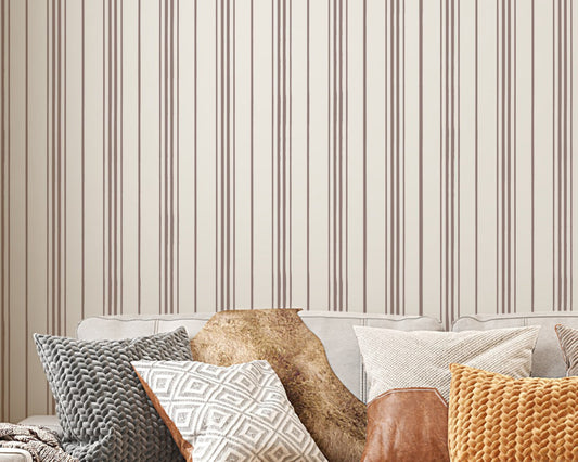 A close-up shot of a beige couch layered with an assortment of textured throw pillows, set against a backdrop of cream-colored wallpaper with vertical brown stripes. The pillows feature various designs, including a grey chunky knit, a white and grey geometric diamond pattern, a smooth brown leather accents, and a bright mustard yellow knit. A fluffy brown faux-fur blanket is also draped over the back of the sofa, creating a warm, cozy interior aesthetic.