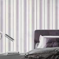 A modern bedroom setting featuring a bed with a deep purple tufted headboard against a white wall with vertical lavender and grey stripes. The bed is neatly styled with grey bedding, a textured dark brown throw blanket, and a mix of lavender and deep purple pillows. To the left, a sleek, minimalist silver desk lamp with a curved arm sits on a dark grey bedside table next to a small stack of books.