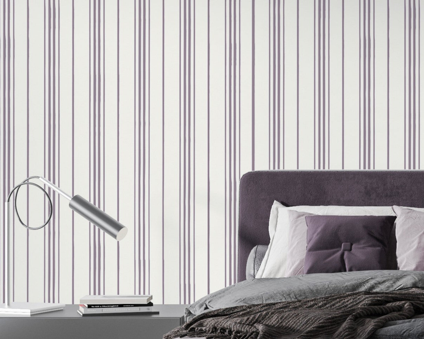 A modern bedroom setting featuring a bed with a deep purple tufted headboard against a white wall with vertical lavender and grey stripes. The bed is neatly styled with grey bedding, a textured dark brown throw blanket, and a mix of lavender and deep purple pillows. To the left, a sleek, minimalist silver desk lamp with a curved arm sits on a dark grey bedside table next to a small stack of books.
