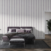 A modern bedroom setting featuring a bed with a deep purple tufted headboard against a white wall with vertical lavender and grey stripes. The bed is neatly styled with grey bedding, a textured dark brown throw blanket, and a mix of lavender and deep purple pillows. To the left, a sleek, minimalist silver desk lamp with a curved arm sits on a dark grey bedside table next to a small stack of books.