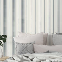 A minimalist bedroom scene featuring soft gray and blush pillows layered on a bed with a textured white blanket. The wall behind the bed is decorated with thin vertical gray stripes on a white background, and a small wooden bedside table holds a white vase and green leafy plant.