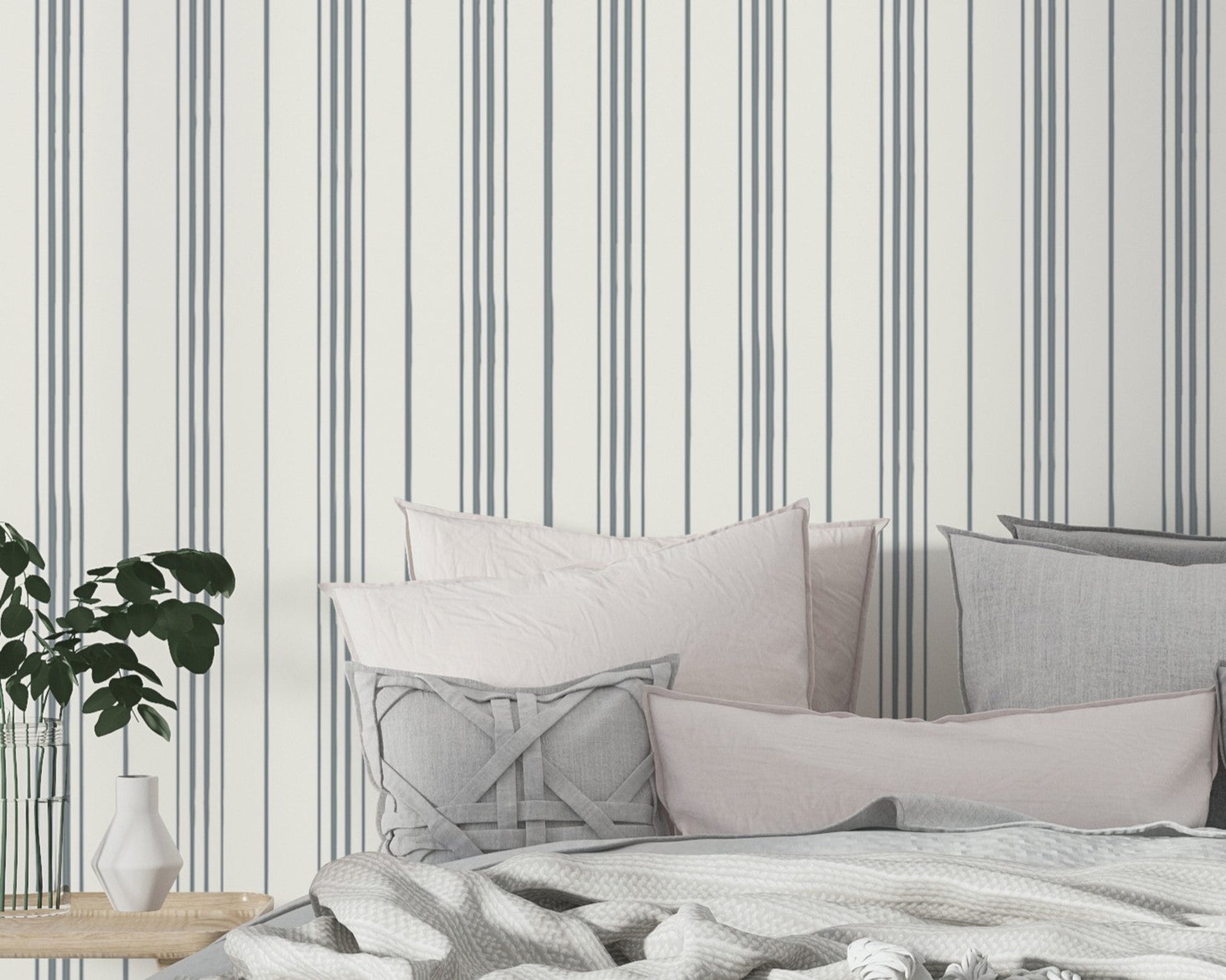 A minimalist bedroom scene featuring soft gray and blush pillows layered on a bed with a textured white blanket. The wall behind the bed is decorated with thin vertical gray stripes on a white background, and a small wooden bedside table holds a white vase and green leafy plant.
