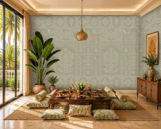 Sunlit tropical dining room with a low wooden table set for a meal on a woven rug, surrounded by floor cushions. Soft sage-green patterned wallpaper covers the walls, while large potted plants, warm brass accents, and a carved pendant light create a relaxed, elegant atmosphere. Floor-to-ceiling glass doors open to a lush palm-filled outdoor view.