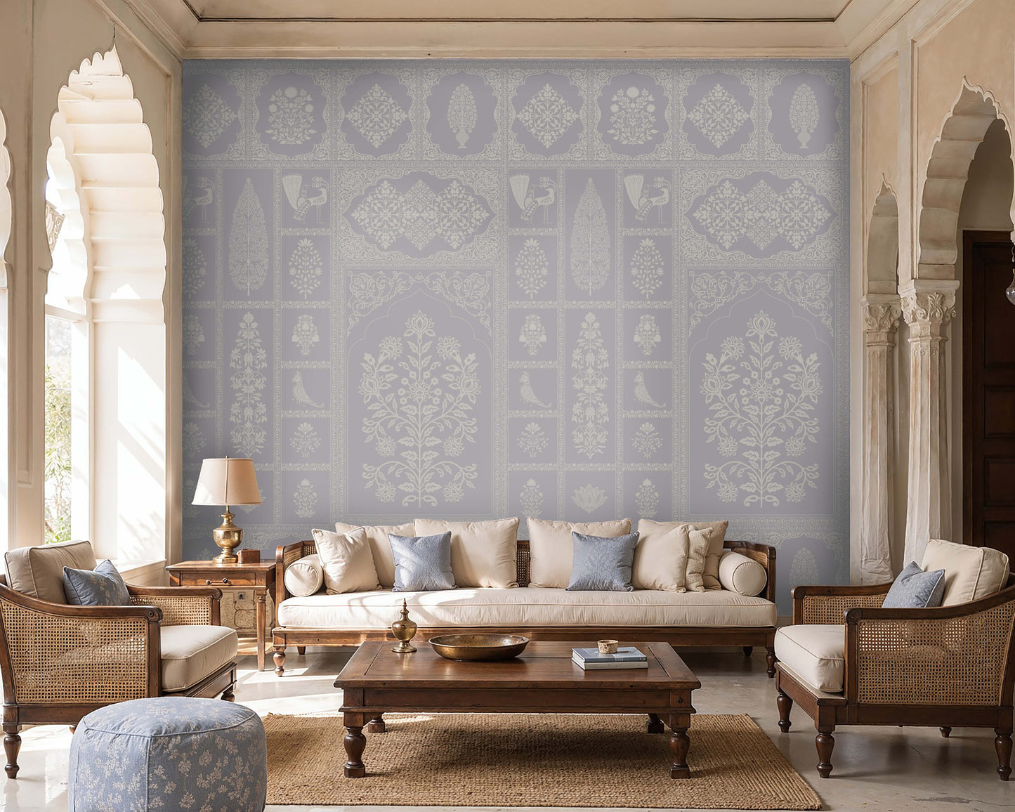 Pastel indigo wallpaper featuring intricate Indian floral motifs, ornamental panels, and traditional decorative elements in soft blue and ivory tones, styled in an elegant living room with wooden cane furniture, neutral upholstery, and arched architectural details.