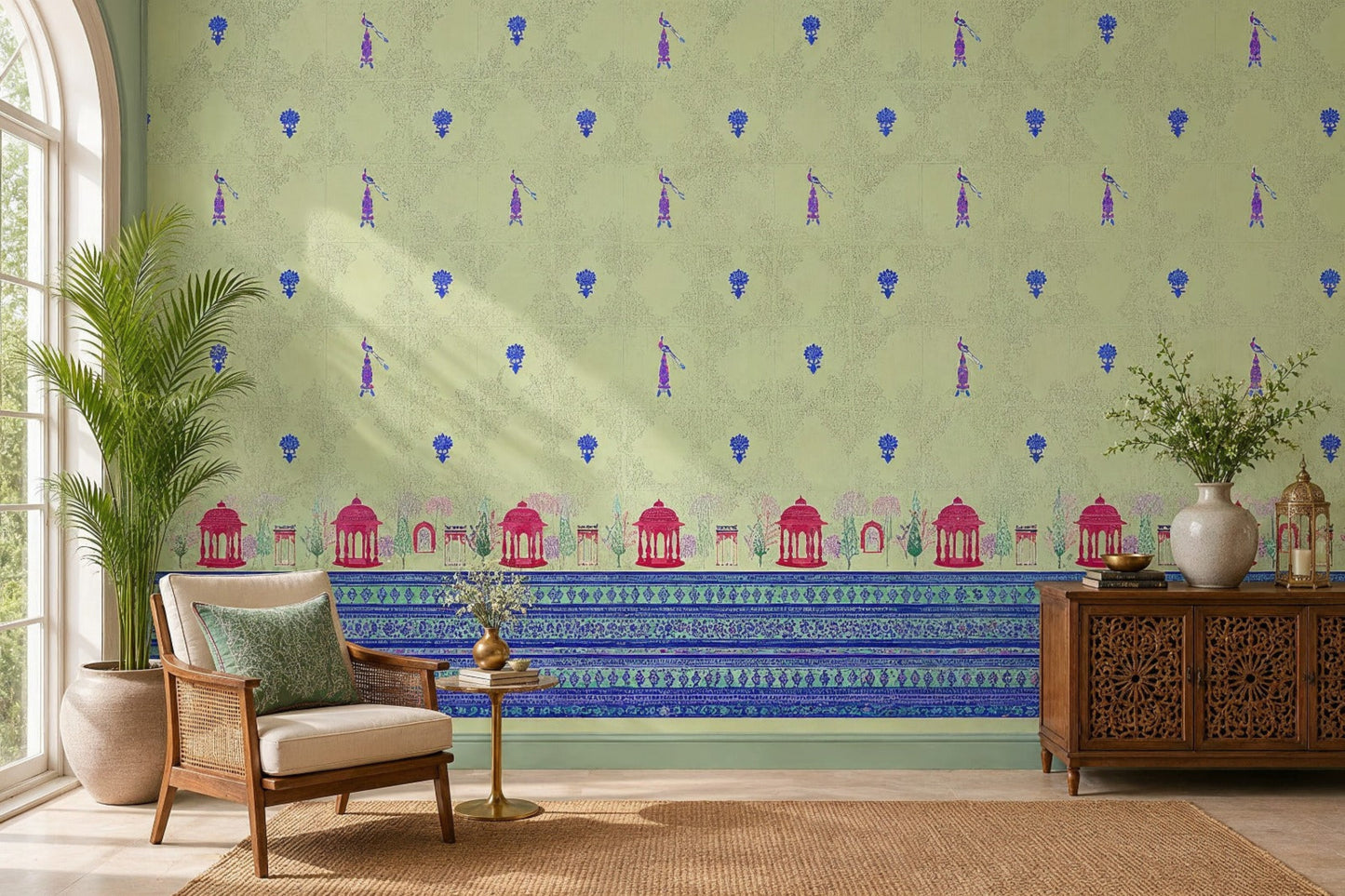 Royal Garden Heritage Wallpaper in Pistachio Green featuring intricate floral motifs, elegant peacock-inspired details, and decorative Mughal garden architecture borders in royal blue and rani pink, styled in a bright living room with cane furniture, carved wooden cabinetry, indoor plants, and natural woven textures.