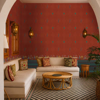 Elegant indoor lounge framed by a large white archway, featuring deep terracotta-red wallpaper with small repeating motifs and a decorative border along the base. The space includes built-in white seating with patterned cushions, a round rattan coffee table, Moroccan-style wall lanterns, and a black-and-white geometric tiled floor. Warm lighting and tropical greenery create a sophisticated, courtyard-inspired atmosphere with Indian and Moroccan design influences.