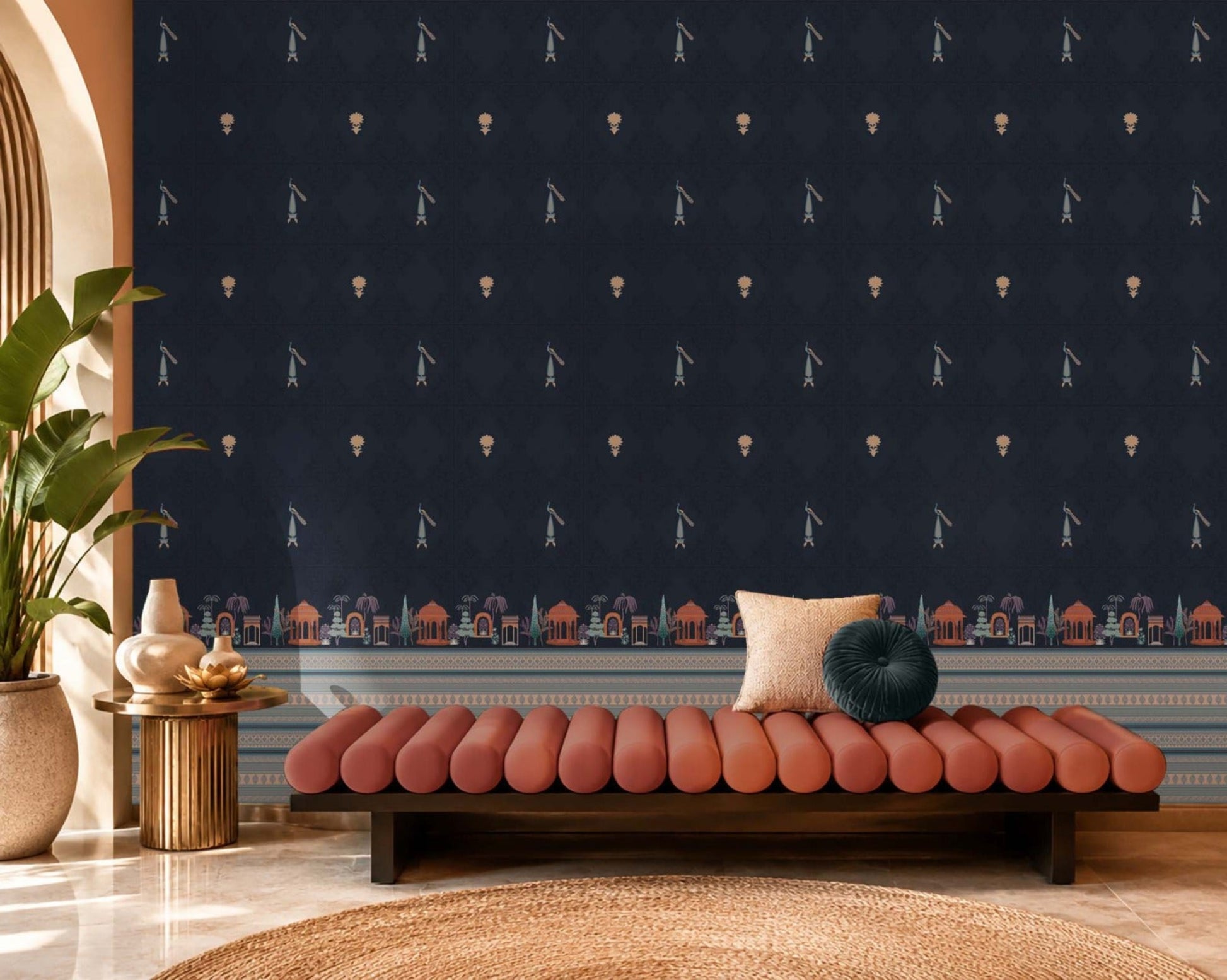 Stylish lounge area featuring a deep navy-blue accent wall decorated with small repeating motifs and an ornate architectural border along the base. The space includes a blush pink upholstered bench with accent cushions, a brass side table with decorative ceramics, and a large potted plant beside an arched window. Warm natural light, soft textures, and rich contrasting colors create an elegant, contemporary interior with Indian-inspired design elements.