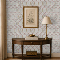 Traditional Indian-inspired wallpaper featuring a delicate trellis pattern with Pichwai-style floral and cow motifs in soft blush and gold tones, displayed in an elegant entryway with a dark wood console table, brass lamp, and framed botanical artwork.
