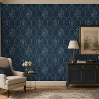 Pichwai-inspired wallpaper in deep navy blue featuring an elegant trellis pattern with traditional floral and cow motifs, styled in a sophisticated living room with a cream armchair, dark blue console cabinet, brass lamp, and classic décor accents.
