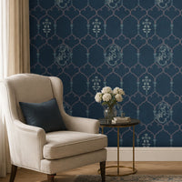Elegant reading nook featuring deep navy-blue wallpaper with an intricate lattice pattern inspired by Indian jaali architecture, accented by delicate floral motifs and stylized face illustrations. A cream upholstered armchair with a navy accent cushion sits beside a round brass side table holding a vase of white flowers and stacked books. Soft natural light and neutral furnishings create a refined, contemporary space with Indian-inspired design elements.
