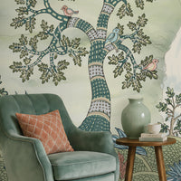 Pastel woodland wall mural featuring hand-painted sage green trees, birds in flight, and deer in a serene forest landscape, styled in a cozy living room with a green armchair and wooden console table.