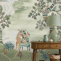 Pastel woodland wall mural featuring hand-painted sage green trees, birds in flight, and deer in a serene forest landscape, styled in a cozy living room with a green armchair and wooden console table.