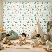 A bright, cheerful children's playroom featuring a nautical-themed wallpaper with a repeating pattern of whales, lighthouses, sailboats, ships, sea turtles, crabs, jellyfish, submarines, anchors, coral, and palm trees in soft pastel shades of teal, coral, mint, and beige. Three young children sit on a round rug playing with wooden blocks, a toy train, and a bead maze. The room includes a canvas play tent, a mint-green bean bag, a wooden rocking horse, open shelving filled with books and toys
