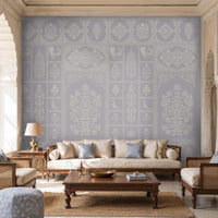 Pastel indigo wallpaper featuring intricate Indian floral motifs, ornamental panels, and traditional decorative elements in soft blue and ivory tones, styled in an elegant living room with wooden cane furniture, neutral upholstery, and arched architectural details.