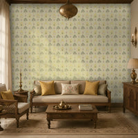 A cozy living room featuring a vintage-inspired wallpaper with a repeating botanical pattern in soft sage green, mustard yellow, dusty pink, and cream on a warm ivory background. The space is furnished with carved wooden seating upholstered in beige fabric, mustard accent cushions, a floral embroidered throw pillow, a wooden coffee table with brass décor, and matching side cabinets. Brass lighting, traditional accessories, and a neutral patterned rug complement the warm, heritage-style interi
