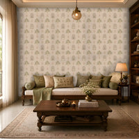 A cozy, elegant living room with a wooden daybed-style sofa topped with olive green and beige embroidered cushions and a matching throw. Behind the sofa, a feature wall is covered in a soft cream wallpaper with a repeating pattern of stylized green trees, arched niches, and delicate pink floral motifs. A dark wood coffee table sits on a light patterned area rug, decorated with books, a brass vase of white flowers, candles, and a small tea set. 