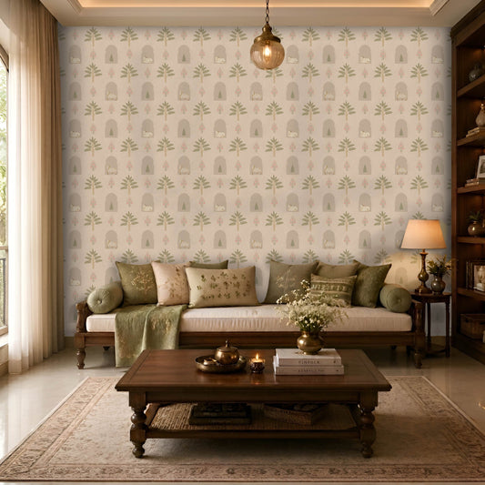 A cozy, elegant living room with a wooden daybed-style sofa topped with olive green and beige embroidered cushions and a matching throw. Behind the sofa, a feature wall is covered in a soft cream wallpaper with a repeating pattern of stylized green trees, arched niches, and delicate pink floral motifs. A dark wood coffee table sits on a light patterned area rug, decorated with books, a brass vase of white flowers, candles, and a small tea set. 