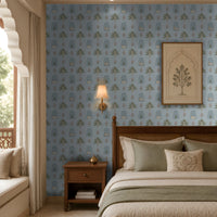 A serene bedroom featuring a light blue wallpaper with a repeating pattern of stylized green trees, arched niches, and delicate pink floral motifs. A wooden bed with a woven cane headboard is dressed in crisp white bedding layered with sage green cushions and a matching throw. Above the bed hangs a framed botanical artwork, while a warm wall sconce and wooden bedside table add soft ambient lighting. 