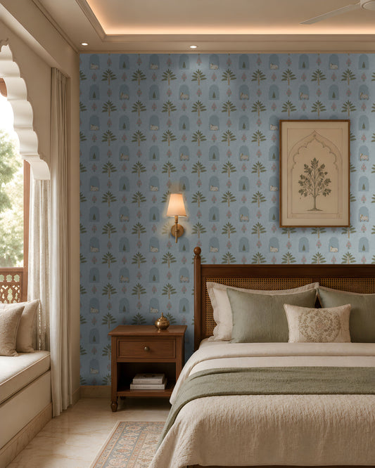 A serene bedroom featuring a light blue wallpaper with a repeating pattern of stylized green trees, arched niches, and delicate pink floral motifs. A wooden bed with a woven cane headboard is dressed in crisp white bedding layered with sage green cushions and a matching throw. Above the bed hangs a framed botanical artwork, while a warm wall sconce and wooden bedside table add soft ambient lighting. 