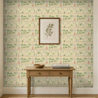 A light-filled entryway features a feature wall covered in soft ivory wallpaper with a repeating Mughal-inspired design of arches, domed pavilions, palm trees, and figures in traditional attire in muted green and blush tones. Centered against the wall is a natural wood console table with two drawers, styled with a wooden bowl and stacked books beneath a framed botanical print. Large windows and neutral trim frame the space, creating a bright, airy setting that highlights the elegant Indian