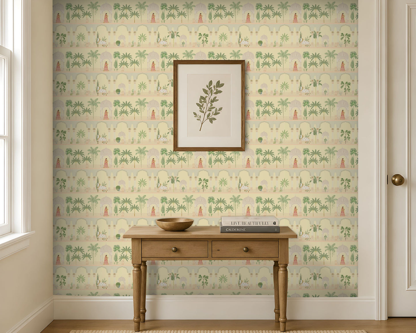 A light-filled entryway features a feature wall covered in soft ivory wallpaper with a repeating Mughal-inspired design of arches, domed pavilions, palm trees, and figures in traditional attire in muted green and blush tones. Centered against the wall is a natural wood console table with two drawers, styled with a wooden bowl and stacked books beneath a framed botanical print. Large windows and neutral trim frame the space, creating a bright, airy setting that highlights the elegant Indian