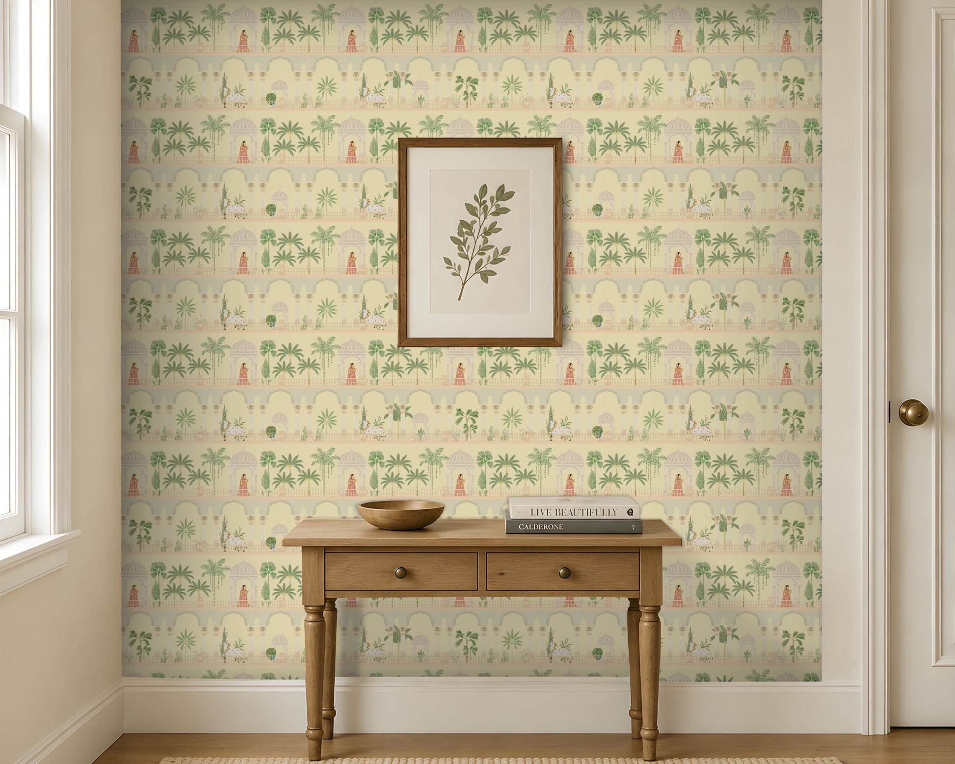 A light-filled entryway features a feature wall covered in soft ivory wallpaper with a repeating Mughal-inspired design of arches, domed pavilions, palm trees, and figures in traditional attire in muted green and blush tones. Centered against the wall is a natural wood console table with two drawers, styled with a wooden bowl and stacked books beneath a framed botanical print. Large windows and neutral trim frame the space, creating a bright, airy setting that highlights the elegant Indian