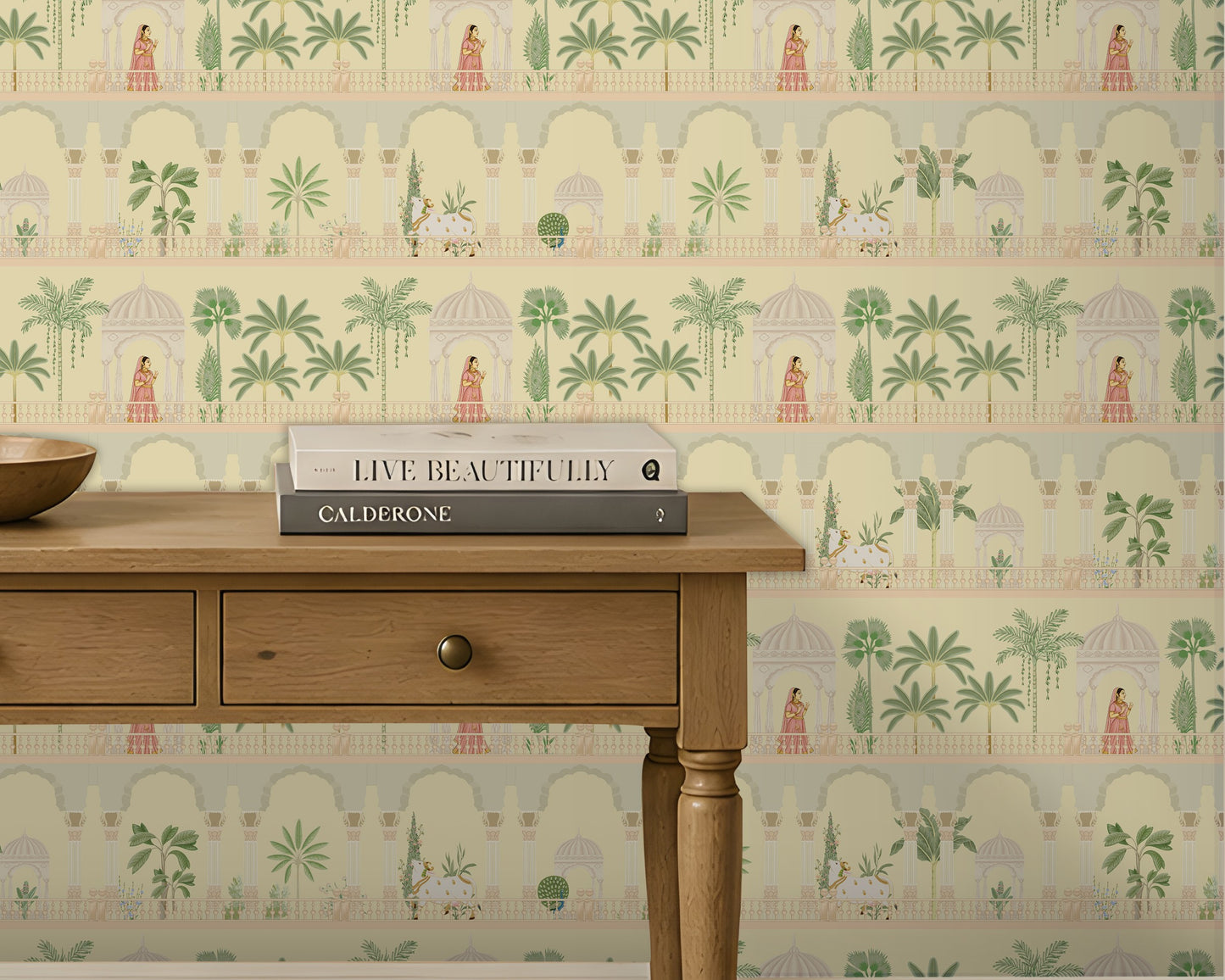 A light-filled entryway features a feature wall covered in soft ivory wallpaper with a repeating Mughal-inspired design of arches, domed pavilions, palm trees, and figures in traditional attire in muted green and blush tones. Centered against the wall is a natural wood console table with two drawers, styled with a wooden bowl and stacked books beneath a framed botanical print. Large windows and neutral trim frame the space, creating a bright, airy setting that highlights the elegant Indian