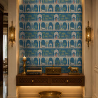 A recessed entryway display features a striking blue-and-white wallpaper with repeating Mughal-inspired arches, domed pavilions, palm trees, elephants, and figures in traditional attire. A dark wood floating console with lattice-front drawers sits below, styled with brass décor, stacked books, a candle, and a decorative animal figurine. Warm recessed and under-cabinet lighting, paired with ornate brass wall sconces, highlights the niche, while a large brass bowl rests on the floor beneath