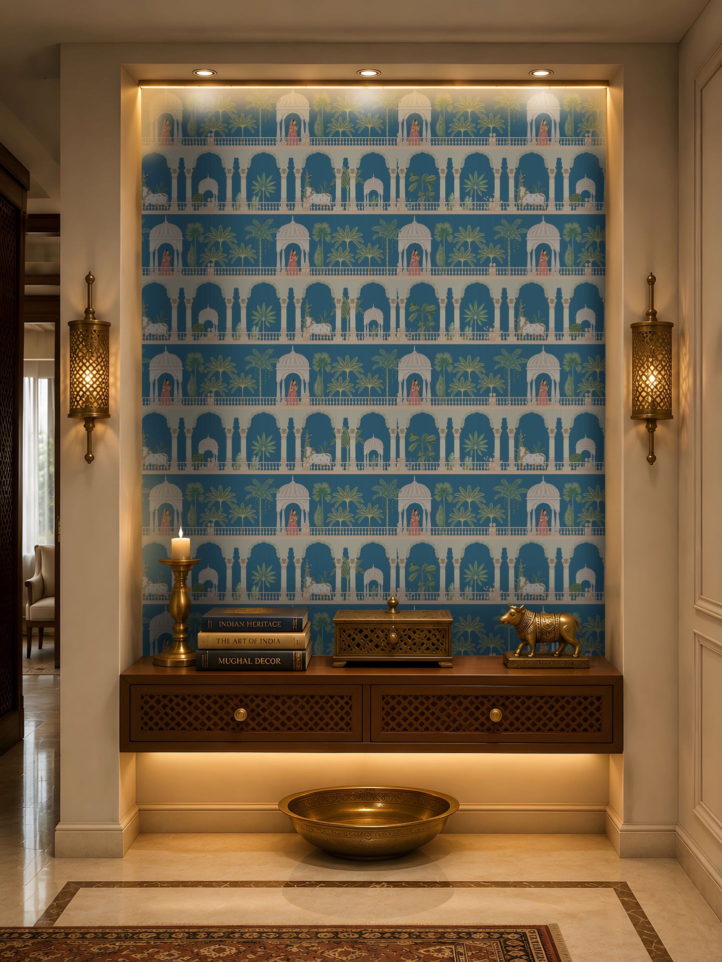 A recessed entryway display features a striking blue-and-white wallpaper with repeating Mughal-inspired arches, domed pavilions, palm trees, elephants, and figures in traditional attire. A dark wood floating console with lattice-front drawers sits below, styled with brass décor, stacked books, a candle, and a decorative animal figurine. Warm recessed and under-cabinet lighting, paired with ornate brass wall sconces, highlights the niche, while a large brass bowl rests on the floor beneath