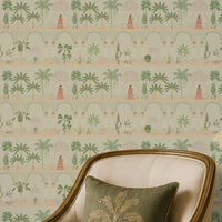 A serene sitting area features a feature wall covered in soft ivory wallpaper with a repeating Mughal-inspired pattern of arches, domed pavilions, palm trees, and figures in traditional attire in muted green and blush tones. In front of the wallpaper, a cream upholstered accent chair with brass trim is paired with a round white-and-gold side table holding a small vase, brass bowl, and stacked books. A ribbed cabinet with a brass table lamp and decorative figurine sits to the side