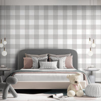 A serene nursery room features a soft gingham check wallpaper in cloud grey.
The design displays a large grid pattern with alternating light grey and white blocks.
In front, a white wooden baby crib filled with plush star and animal toys is centered.
The calm and peaceful interior layout is nicely completed by a light grey rug on the floor.