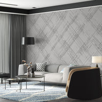 A modern interior bedroom features a textured crosshatch wallpaper in a rhino grey theme.
The fine fabric-look pattern displays intersecting white sketch lines over a cool grey background.
In front, a bed with clean white sheets and dark charcoal pillows sits next to a wooden table.
The bright, contemporary layout is beautifully completed by a sleek black metal reading lamp on the left.