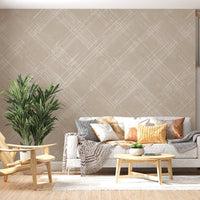 A bright living room features a textured crosshatch wallpaper in a warm beige theme.
The fabric-look pattern displays intersecting white sketch lines angled dynamically over a sandy beige backdrop.
In front, a cozy white sofa with colourful textured pillows sits next to a light wood lounge chair.
The sunny, welcoming interior space is beautifully completed by a tall, leafy green potted plant.