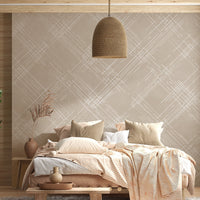 A bright living room features a textured crosshatch wallpaper in a warm beige theme.
The fabric-look pattern displays intersecting white sketch lines angled dynamically over a sandy beige backdrop.
In front, a cozy white sofa with colourful textured pillows sits next to a light wood lounge chair.
The sunny, welcoming interior space is beautifully completed by a tall, leafy green potted plant.