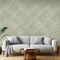 A contemporary living room features a textured crosshatch wallpaper in an olive grey theme.
The fabric-look pattern displays intersecting white sketch lines layered across a muted olive green backdrop.
In front, a comfortable white sofa with grey throw pillows sits behind a round amber glass coffee table.
The calm and stylish interior space is completed by a large leafy green potted plant on the left.