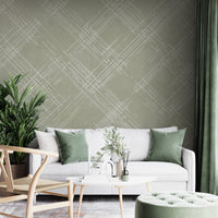 A contemporary living room features a textured crosshatch wallpaper in an olive grey theme.
The fabric-look pattern displays intersecting white sketch lines layered across a muted olive green backdrop.
In front, a comfortable white sofa with grey throw pillows sits behind a round amber glass coffee table.
The calm and stylish interior space is completed by a large leafy green potted plant on the left.