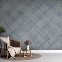 A modern interior features a textured crosshatch wallpaper in a steel grey theme.
The fabric-look pattern displays intersecting dark blue-grey sketch lines over a medium grey background.
In front, a comfortable light beige armchair with a blanket sits on a round woven rug.
The simple, contemporary layout is completed by a small wooden side table holding flowers.