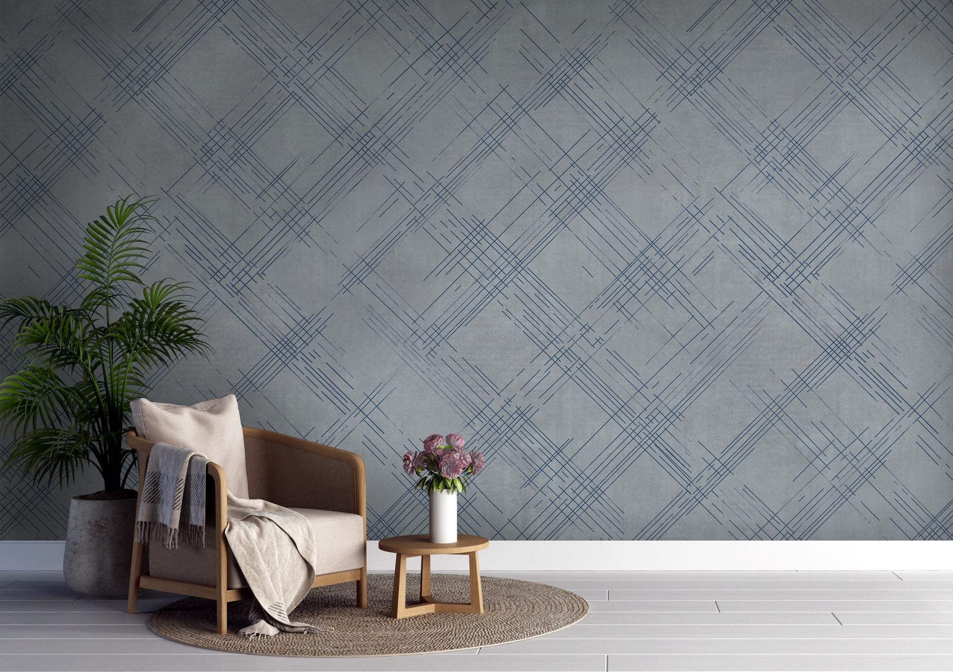 A modern interior features a textured crosshatch wallpaper in a steel grey theme.
The fabric-look pattern displays intersecting dark blue-grey sketch lines over a medium grey background.
In front, a comfortable light beige armchair with a blanket sits on a round woven rug.
The simple, contemporary layout is completed by a small wooden side table holding flowers.