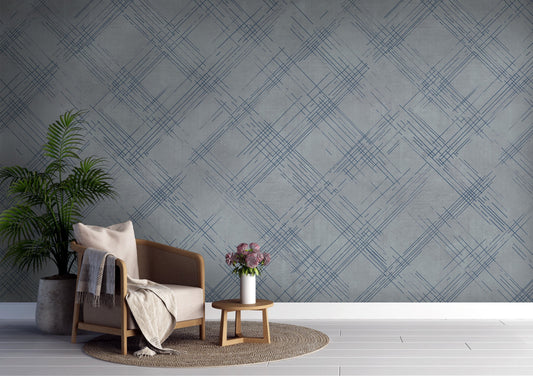 A modern interior features a textured crosshatch wallpaper in a steel grey theme.
The fabric-look pattern displays intersecting dark blue-grey sketch lines over a medium grey background.
In front, a comfortable light beige armchair with a blanket sits on a round woven rug.
The simple, contemporary layout is completed by a small wooden side table holding flowers.