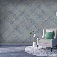 A modern interior features a textured crosshatch wallpaper in a steel grey theme.
The fabric-look pattern displays intersecting dark blue-grey sketch lines over a medium grey background.
In front, a comfortable light beige armchair with a blanket sits on a round woven rug.
The simple, contemporary layout is completed by a small wooden side table holding flowers.