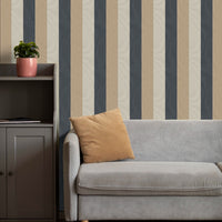 A cozy living room setup features a bold striped wallpaper with a fabric-textured look.
The design displays alternating thick vertical bands of charcoal grey, warm beige, and light cream.
In front, a comfortable light grey fabric sofa with a tan accent pillow is positioned on the right.
The modern interior space is completed by a dark grey wooden storage cabinet placed on the left side.