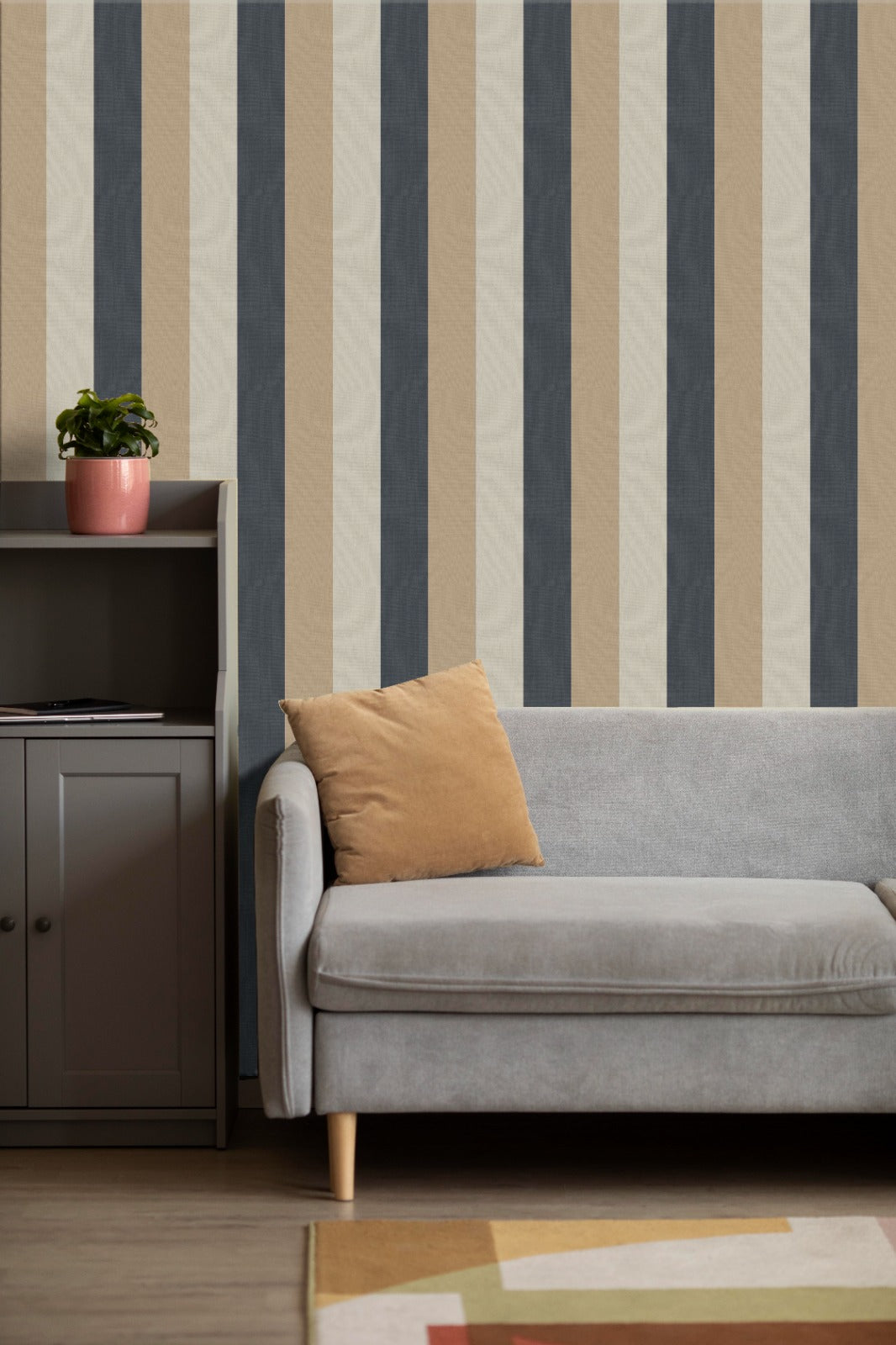 A cozy living room setup features a bold striped wallpaper with a fabric-textured look.
The design displays alternating thick vertical bands of charcoal grey, warm beige, and light cream.
In front, a comfortable light grey fabric sofa with a tan accent pillow is positioned on the right.
The modern interior space is completed by a dark grey wooden storage cabinet placed on the left side.