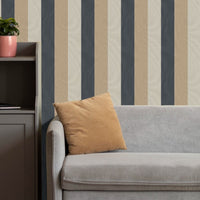 A cozy living room setup features a bold striped wallpaper with a fabric-textured look.
The design displays alternating thick vertical bands of charcoal grey, warm beige, and light cream.
In front, a comfortable light grey fabric sofa with a tan accent pillow is positioned on the right.
The modern interior space is completed by a dark grey wooden storage cabinet placed on the left side.