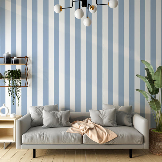 A bright living room features a striped wallpaper pattern.
The wallpaper colors are soft sky blue and crisp white.
The design has clean, evenly spaced vertical stripes across the wall.
A light grey sofa with several matching pillows sits in front.
A pale peach blanket is draped casually over the couch cushions.
The look is finished with a large green plant on the right.