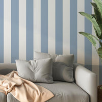 A bright living room features a striped wallpaper pattern.
The wallpaper colors are soft sky blue and crisp white.
The design has clean, evenly spaced vertical stripes across the wall.
A light grey sofa with several matching pillows sits in front.
A pale peach blanket is draped casually over the couch cushions.
The look is finished with a large green plant on the right.
