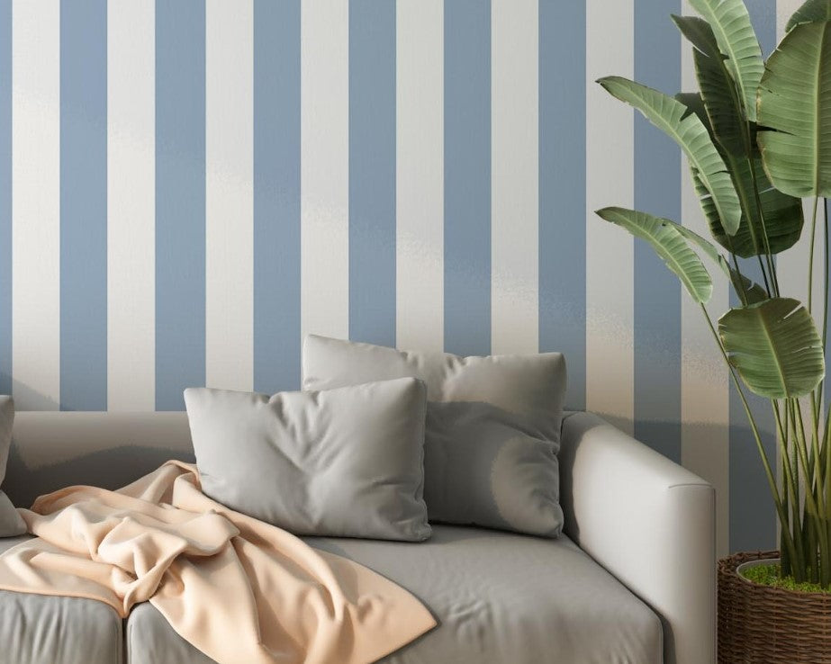 A bright living room features a striped wallpaper pattern.
The wallpaper colors are soft sky blue and crisp white.
The design has clean, evenly spaced vertical stripes across the wall.
A light grey sofa with several matching pillows sits in front.
A pale peach blanket is draped casually over the couch cushions.
The look is finished with a large green plant on the right.