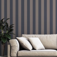 A modern living room features a dark navy blue and muted grey-brown striped wallpaper.
The design showcases clean, evenly spaced vertical stripes filling the entire back wall.
In front, a beige fabric sofa with decorative pillows rests on a chevron pattern rug.
The contemporary, stylish space is finished with a large potted green plant on the left.